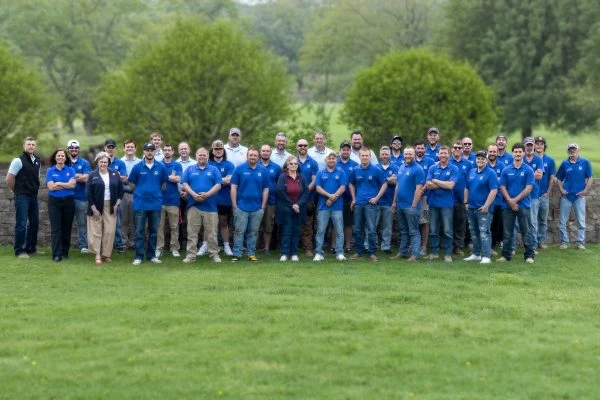 Large group image of the entire company team all wearing blue shirts standing in a field with trees in the background