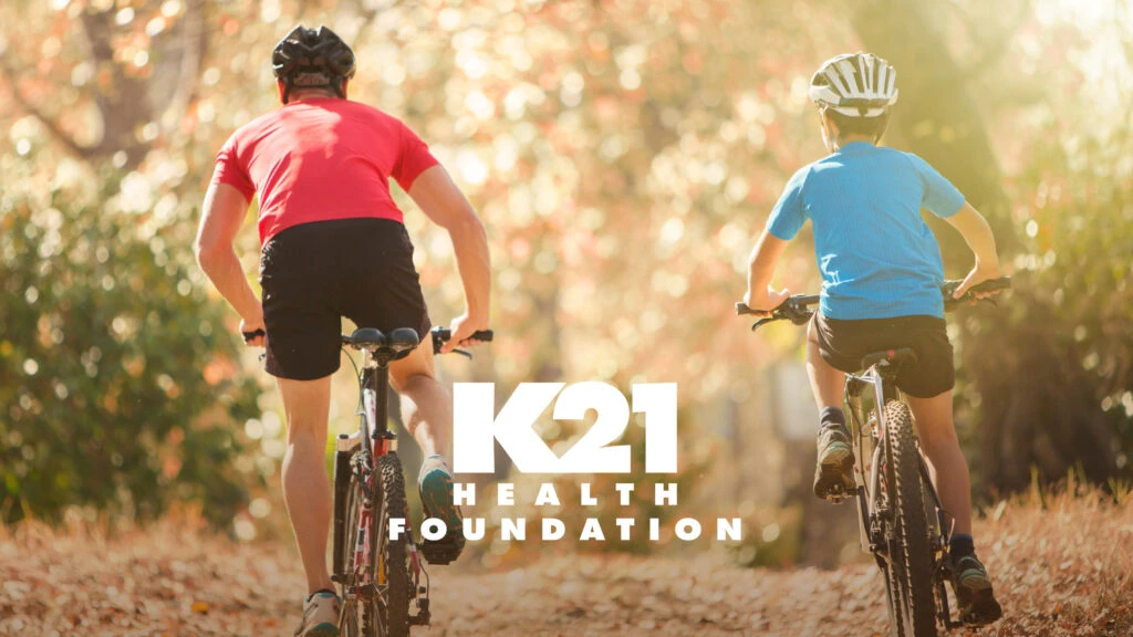 Man and boy riding mountain bikes on a trail with K21 Health Foundation Logo