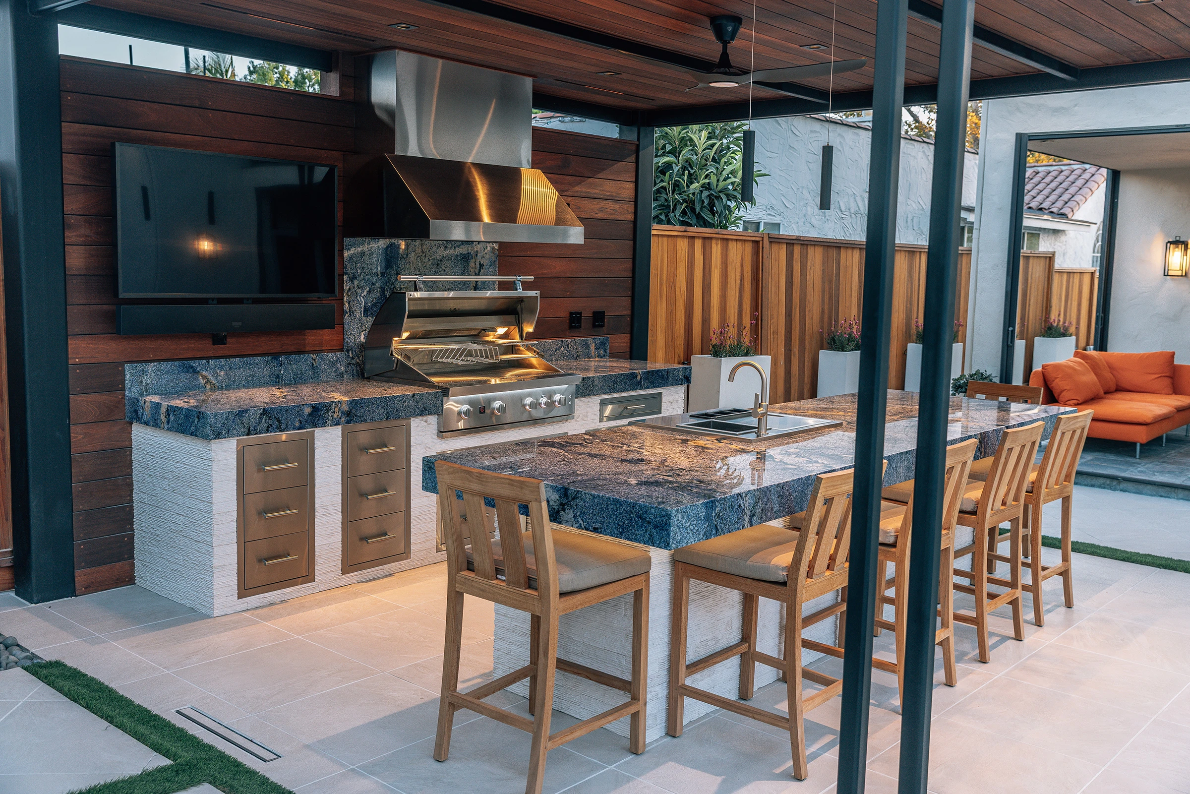 Outdoor kitchen with stainless steel vent hood duct cover over a Summerset grill, stone-top island, and wood cabinet drawers under a covered patio.